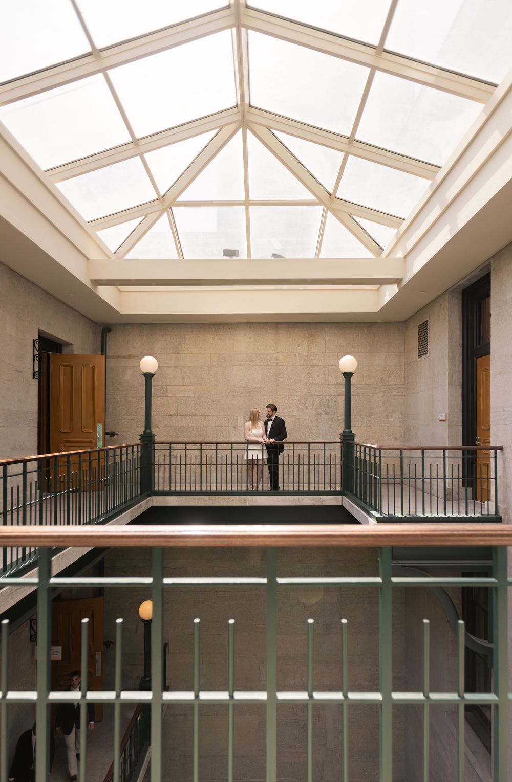 Couple standing on an elegant indoor balcony beneath a large skylight in a classic building atrium.