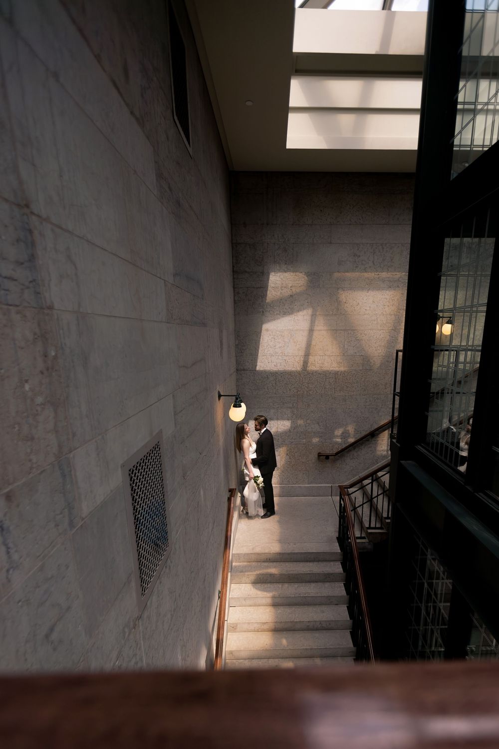 Bride and groom share a romantic moment on elegant stone staircase bathed in natural light from above.