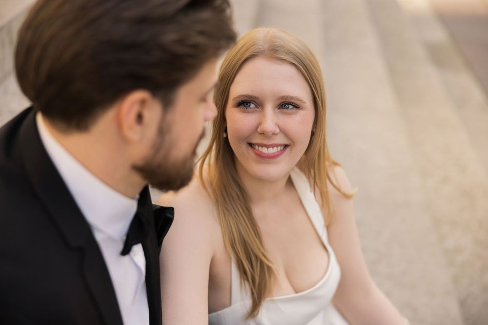A smiling bride in a white dress gazes at a groom in a black tuxedo during an outdoor wedding photo session.
