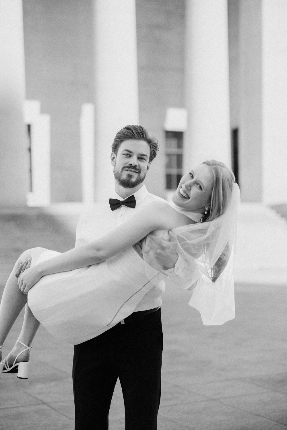 Groom carrying bride in white dress and veil in black and white wedding portrait near classical columns.