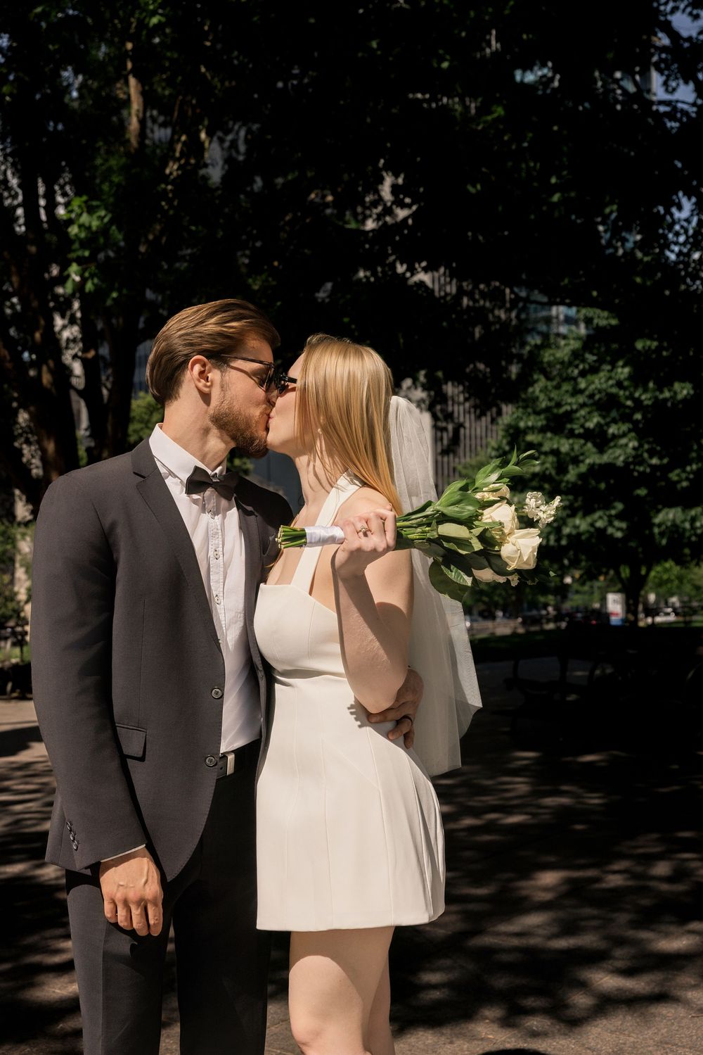 Newlyweds kissing outdoors, bride in short white dress holding tulip bouquet, groom in dark suit with bow tie.