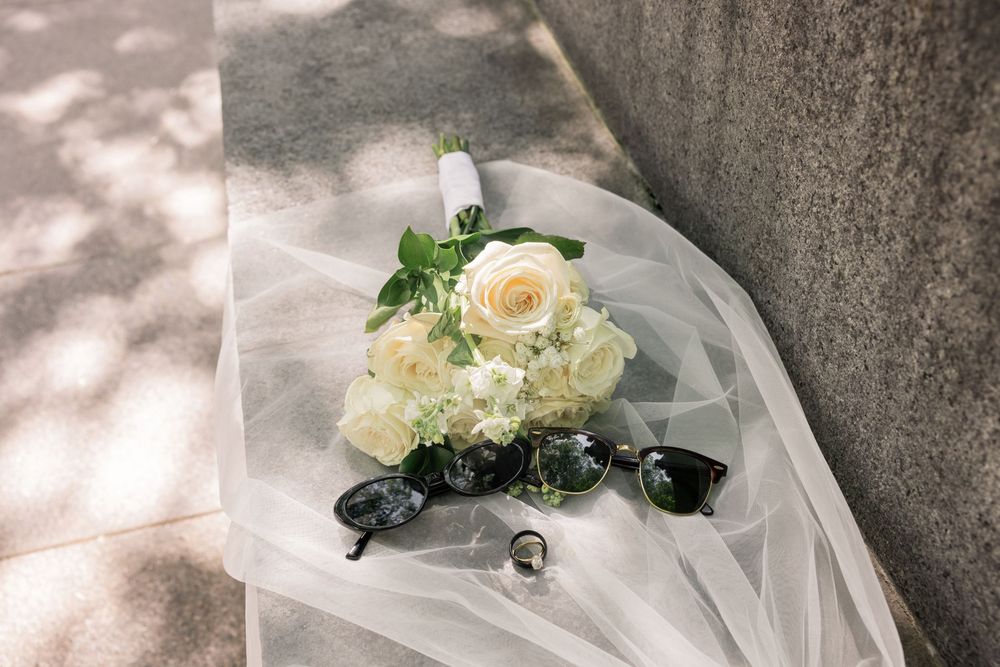 White rose wedding bouquet with baby's breath on tulle veil beside sunglasses and ring on stone surface.