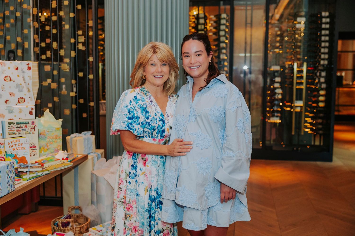 Two women smiling together at a baby shower, surrounded by gifts and decorations in an elegant venue.
