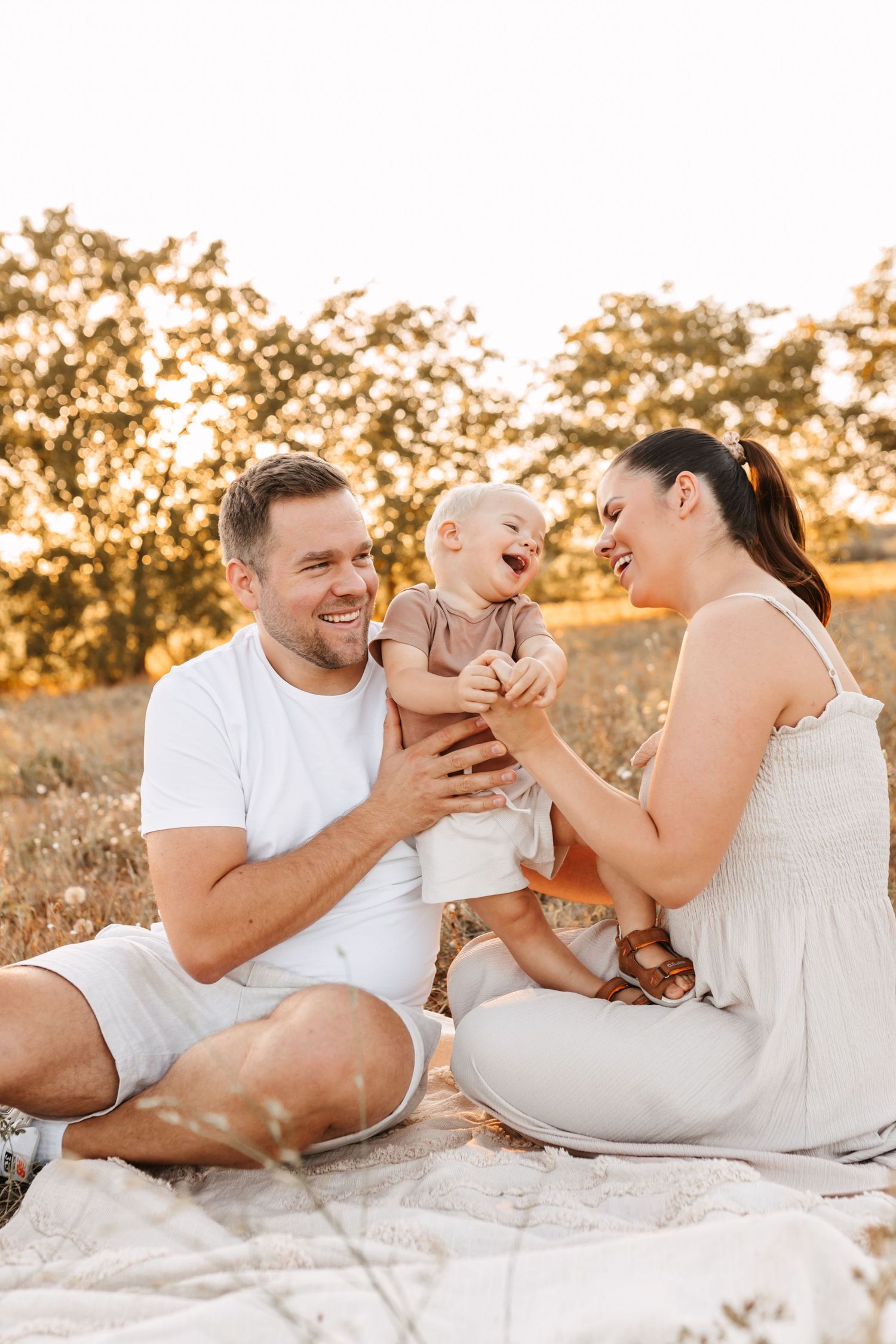 Boldog családi pillanatok. Happy family with laughing baby sitting on blanket in golden sunlit field, parents smiling and playing outdoors.