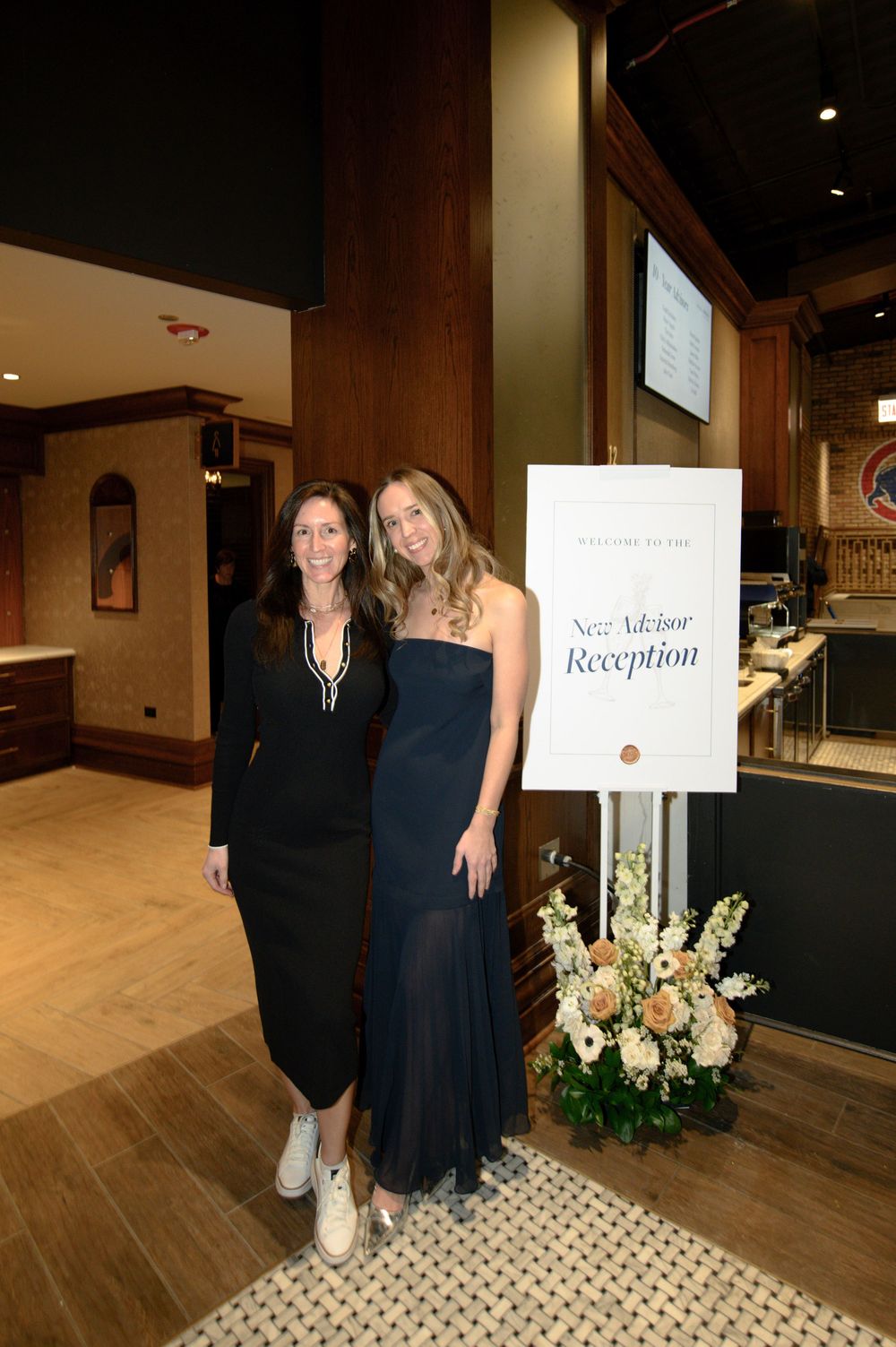 Two women in black dresses pose together at a New Advisor Reception event with floral arrangements nearby.