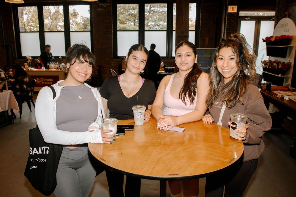 Four young women smiling together at a round wooden table inside a cozy restaurant cafe setting.