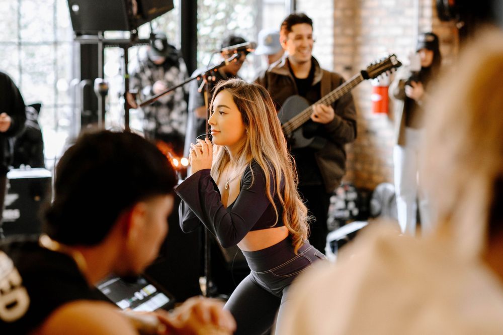 Female singer performing on stage with guitarist and band members in a modern indoor venue.