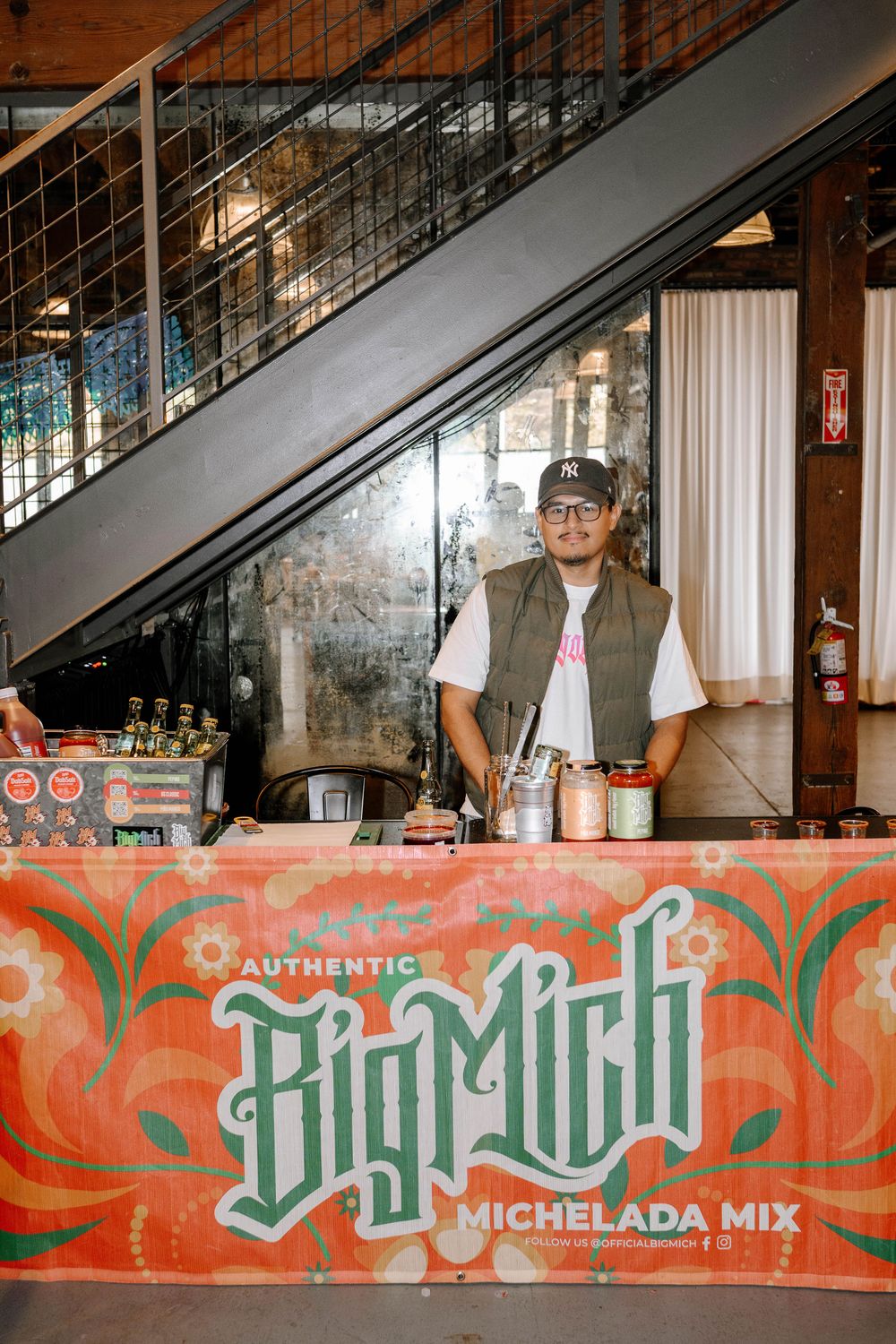 Man standing behind a Big Mich Michelada Mix booth at an indoor event venue with stairs in background.