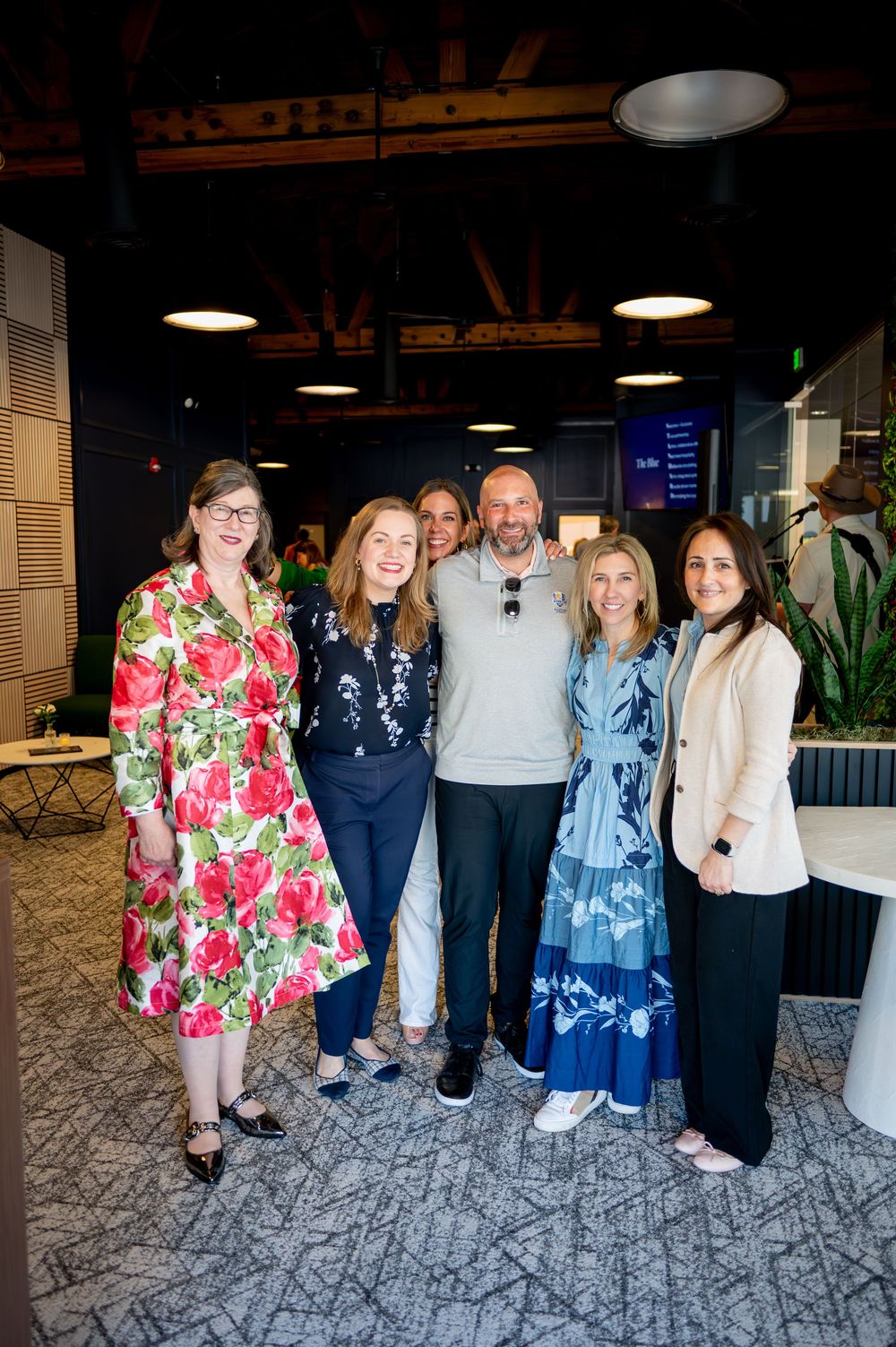 Six professionals smile together in a modern office space, dressed in colorful attire at a corporate networking event.