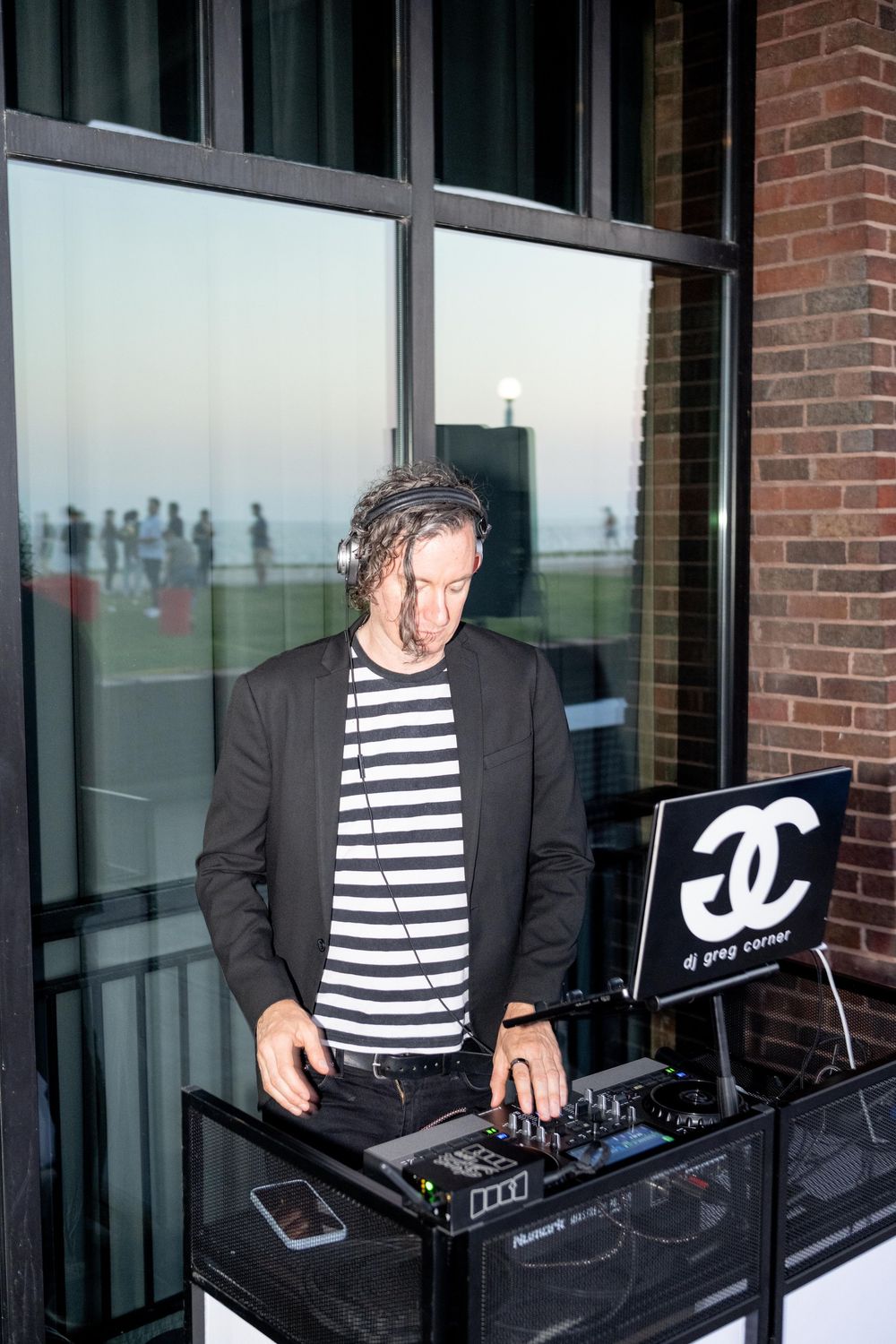 DJ Greg Corner performs at an outdoor event, wearing headphones and a striped shirt behind a Chanel-branded DJ booth.