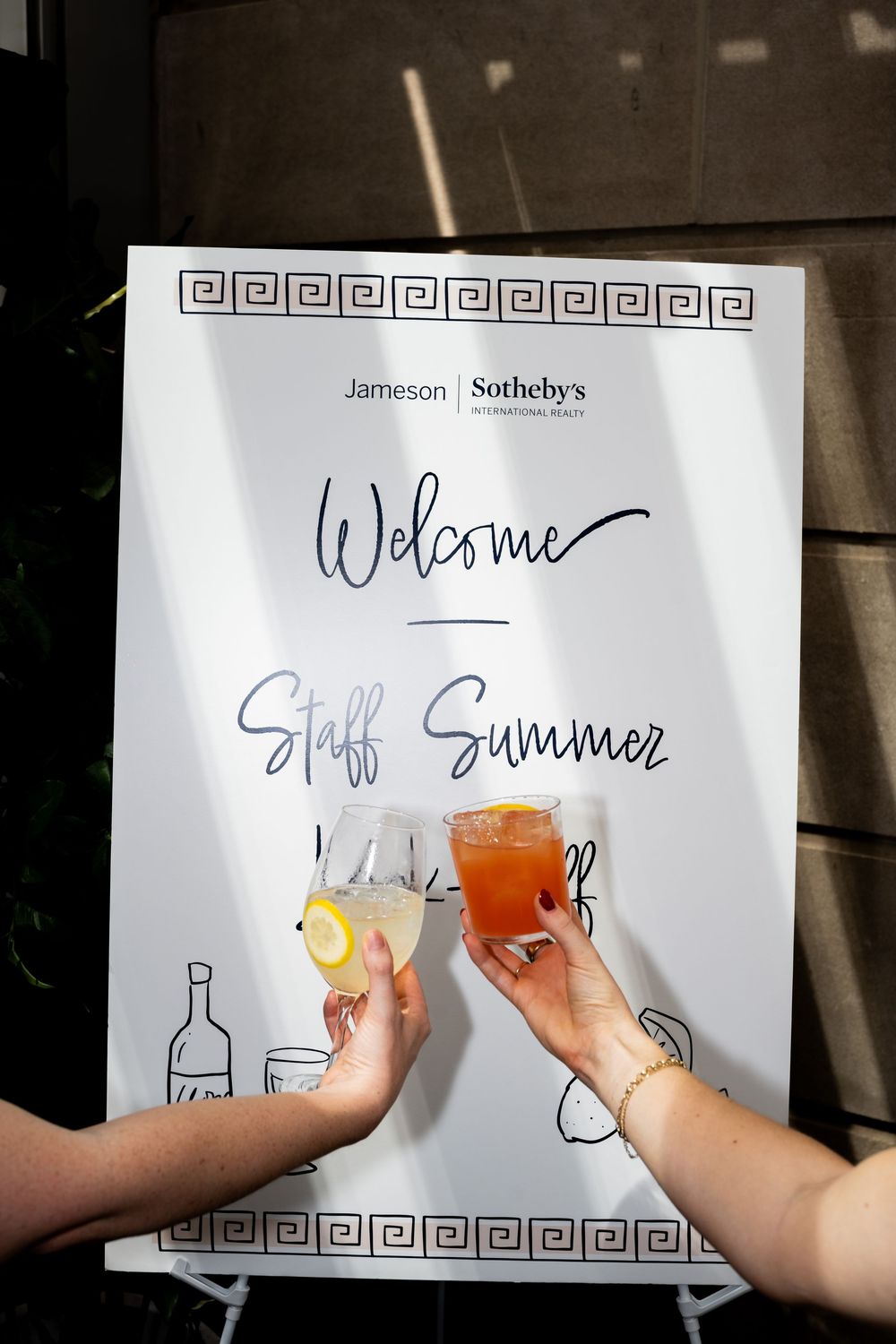 Two hands toasting drinks in front of a Jameson Sotheby's Welcome Staff Summer event sign.