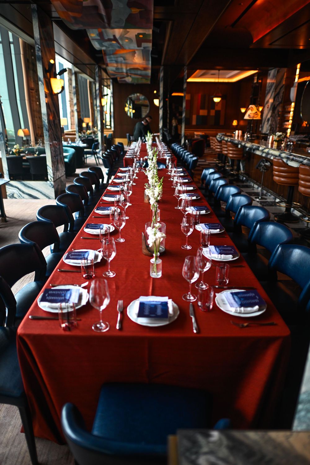 Elegant long dining table with red tablecloth, blue chairs, wine glasses, and place settings in upscale restaurant.