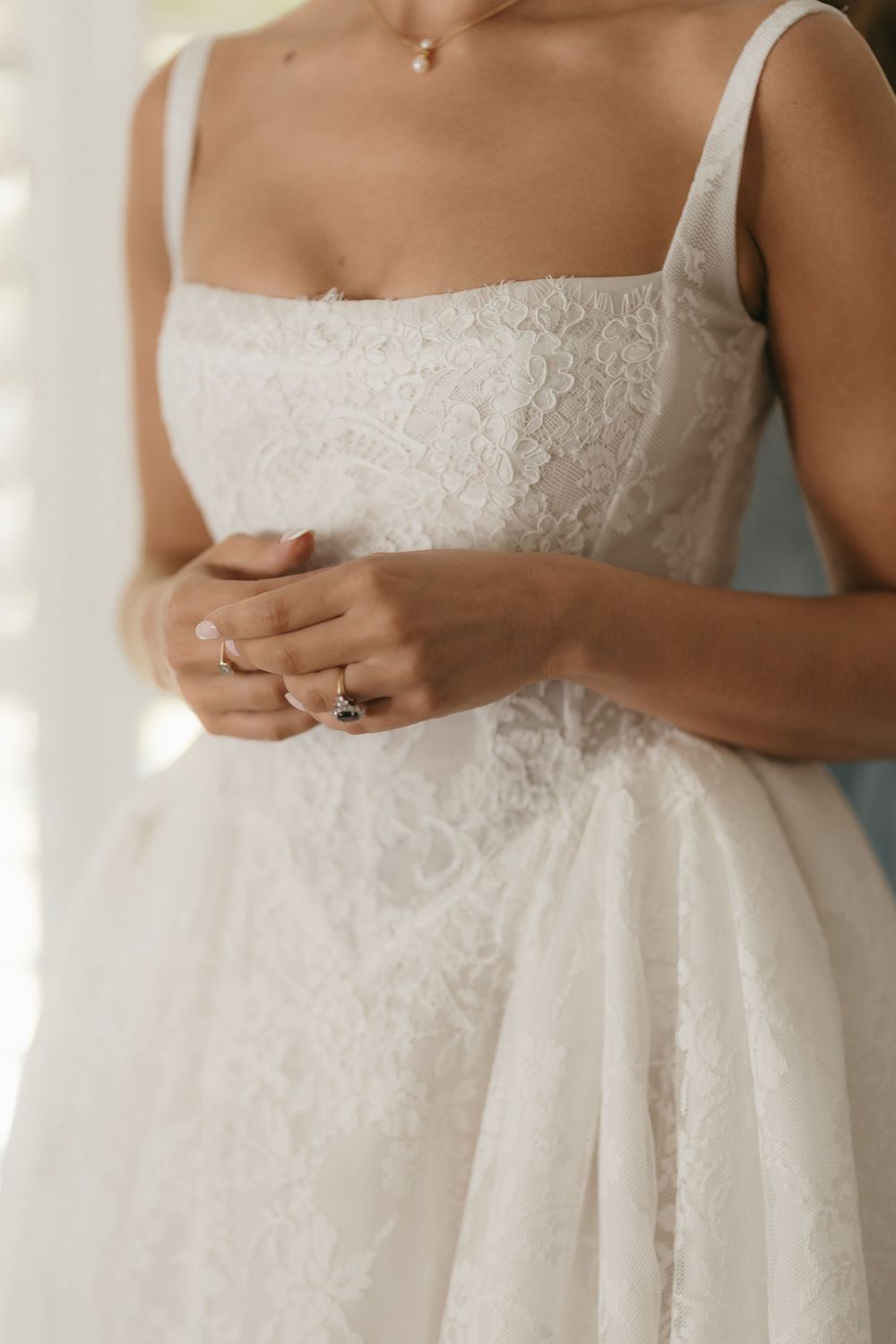 Bride in white lace square-neck wedding dress with gold rings, clasping hands gently at waist.