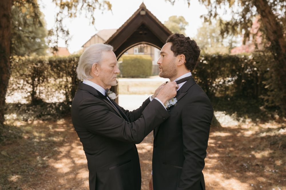 An older man adjusts the bow tie of a young groom in dark suits outdoors before a wedding ceremony.