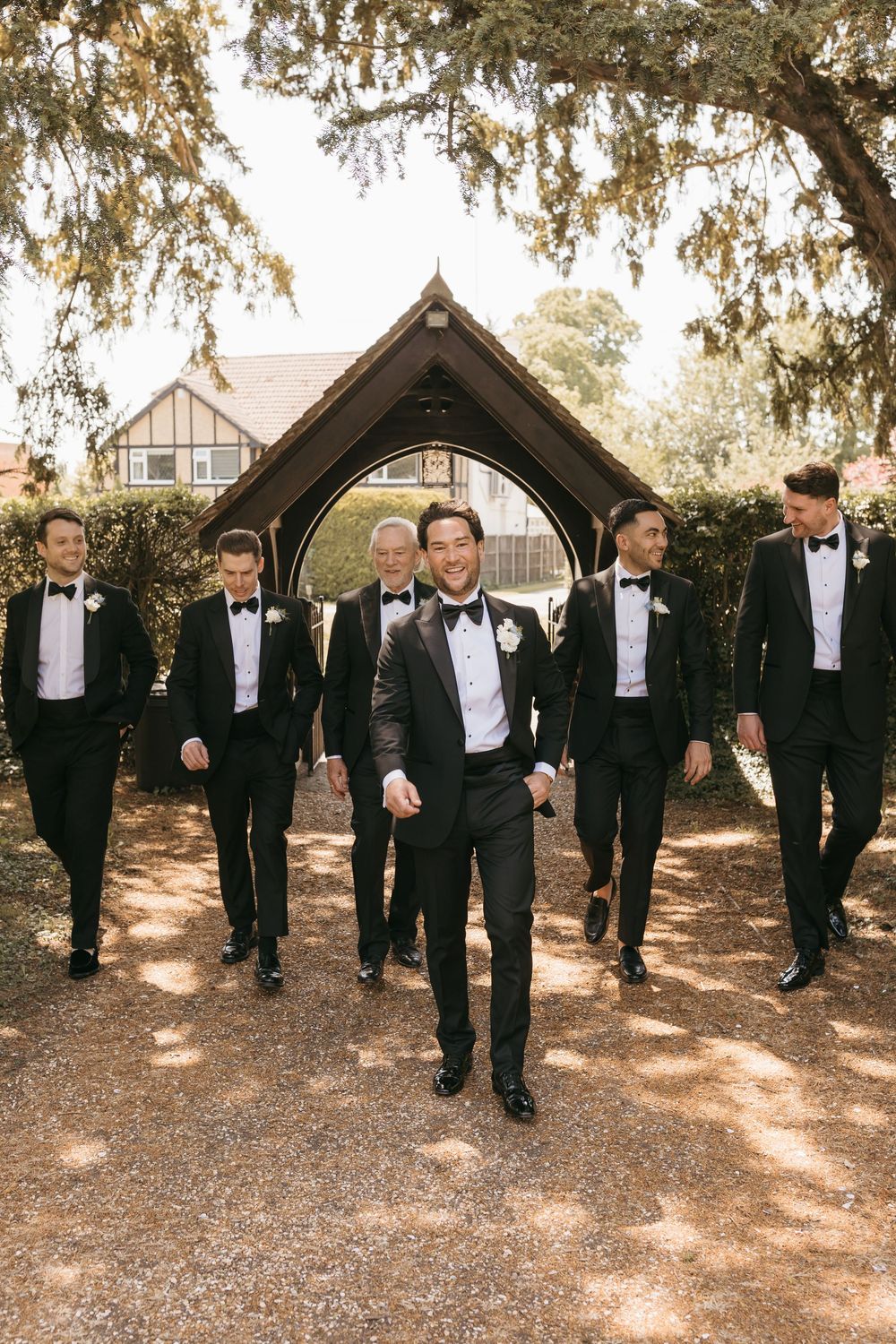 Groom leads groomsmen in black tuxedos walking through an arched garden gate at an outdoor wedding venue.