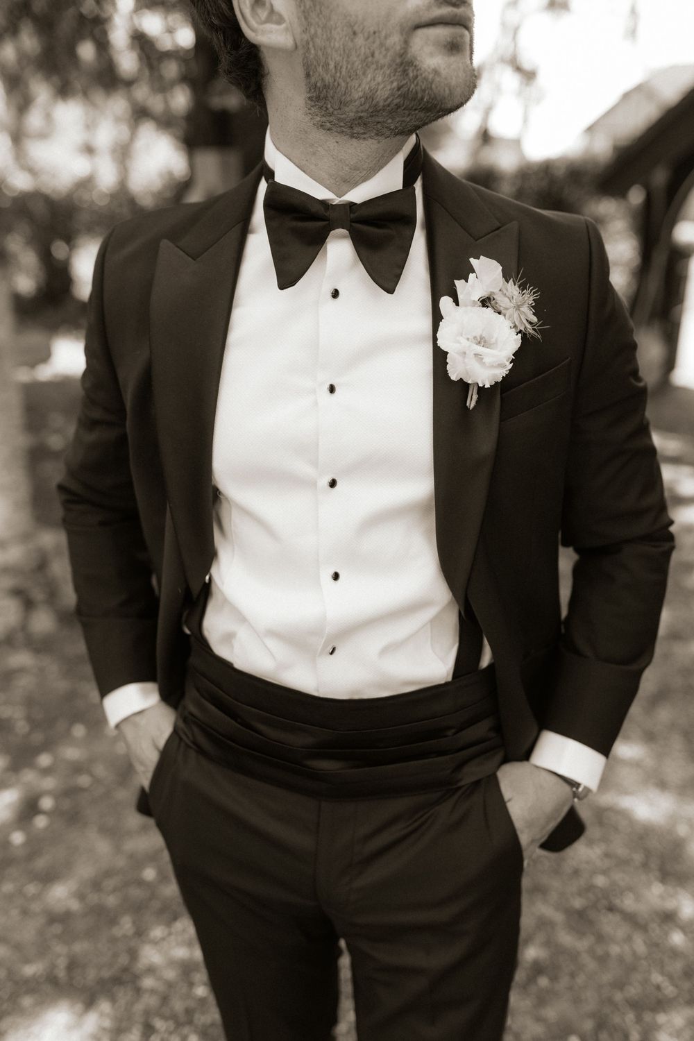 Groom in black tuxedo with bow tie and floral boutonniere, hands in pockets, sepia toned wedding photo.