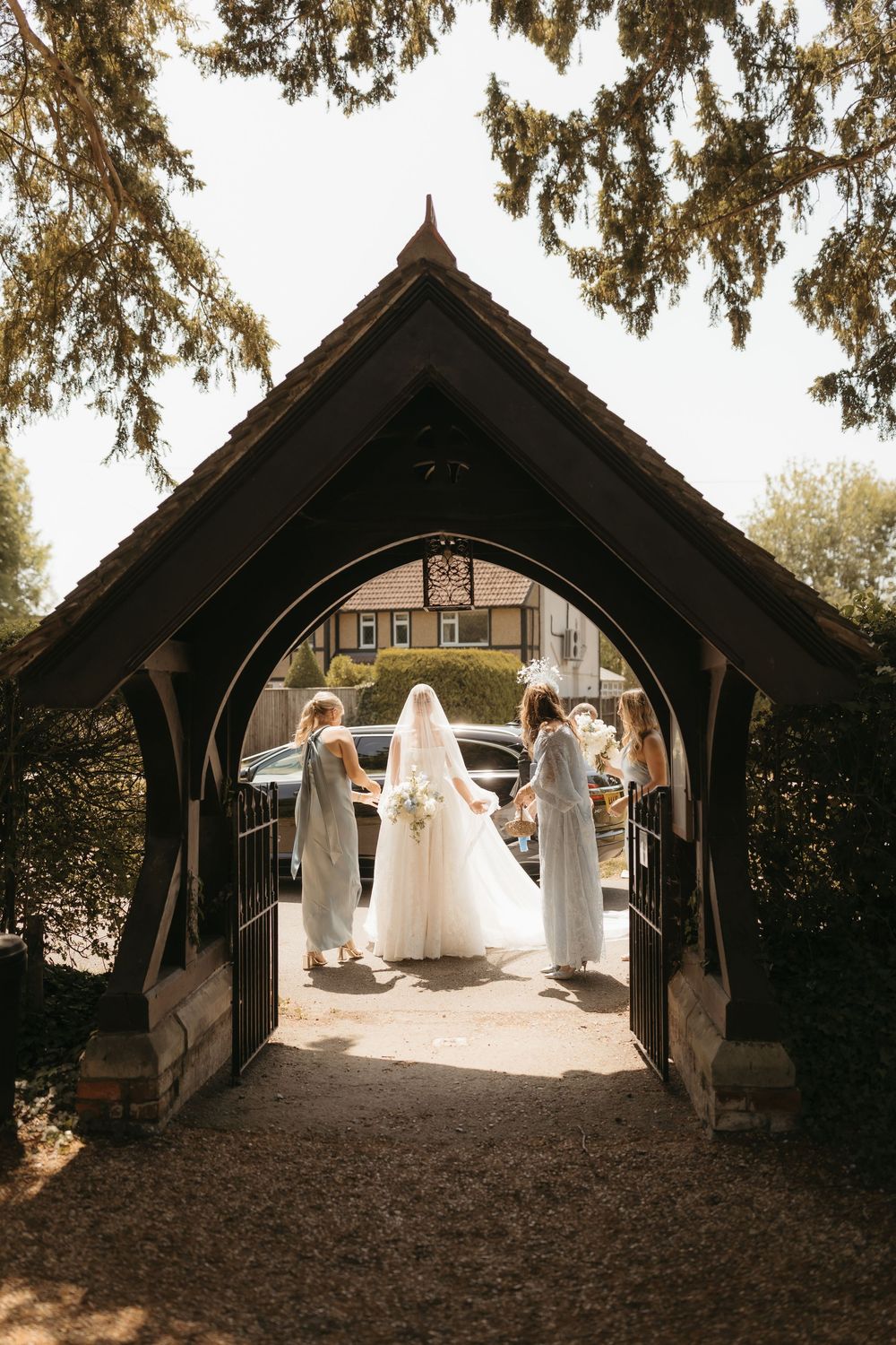 Bride in white gown and veil stands with bridesmaids in gray dresses under a wooden arch gate.