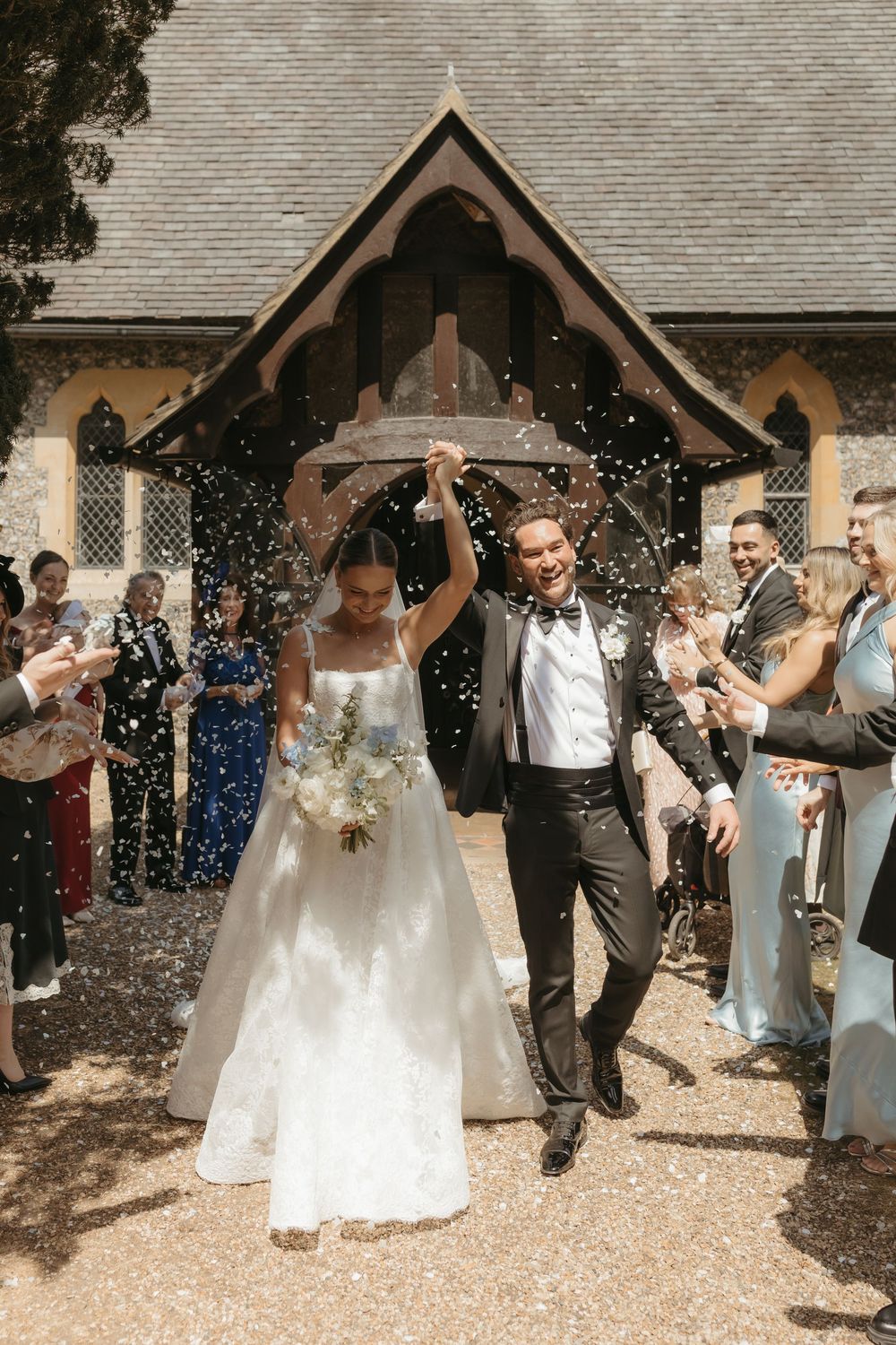 Bride and groom exit church amid confetti shower, guests celebrating their wedding ceremony outdoors.