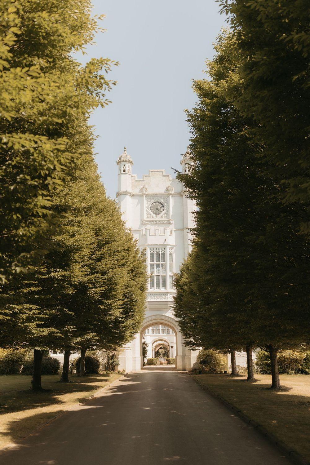 Tree-lined driveway leads to elegant white Gothic manor house under clear blue sky.