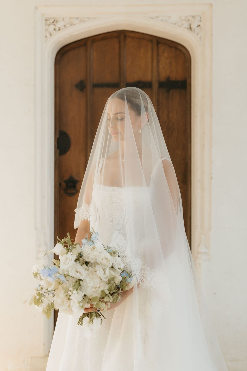 Elegant bride wearing a cathedral veil holding a white and blue floral bouquet standing before a wooden church door.