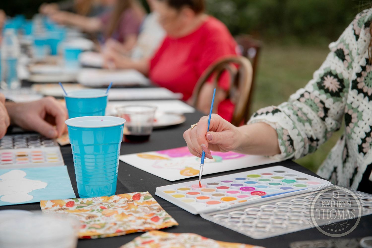 Group of people dining together at long outdoor tables on a grassy field during a warm summer evening gathering.
