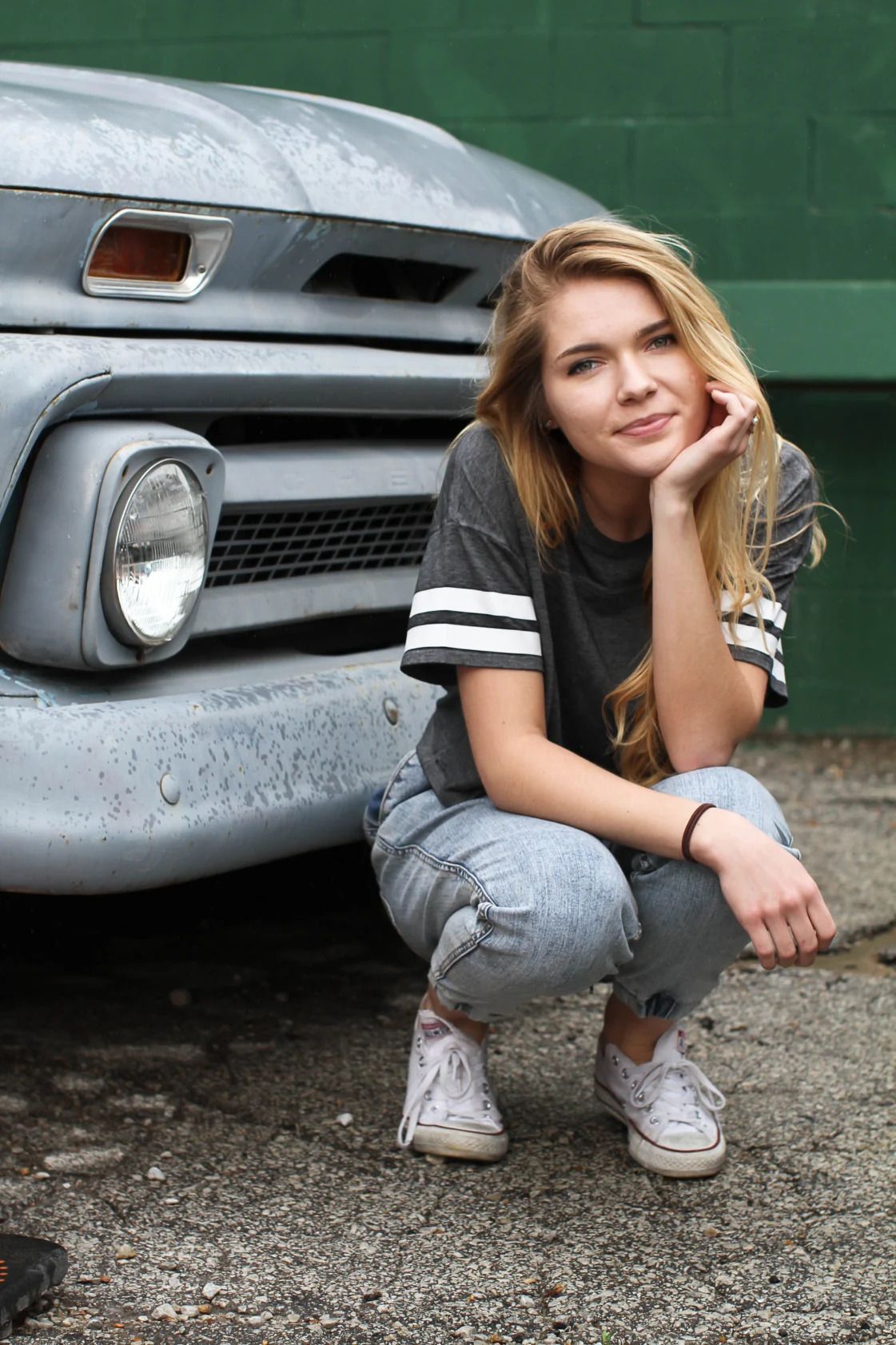 A young woman in jeans & a t-shirt crouches next to an old car in front of a green wall and smiles softly with her chin resting in her hand.