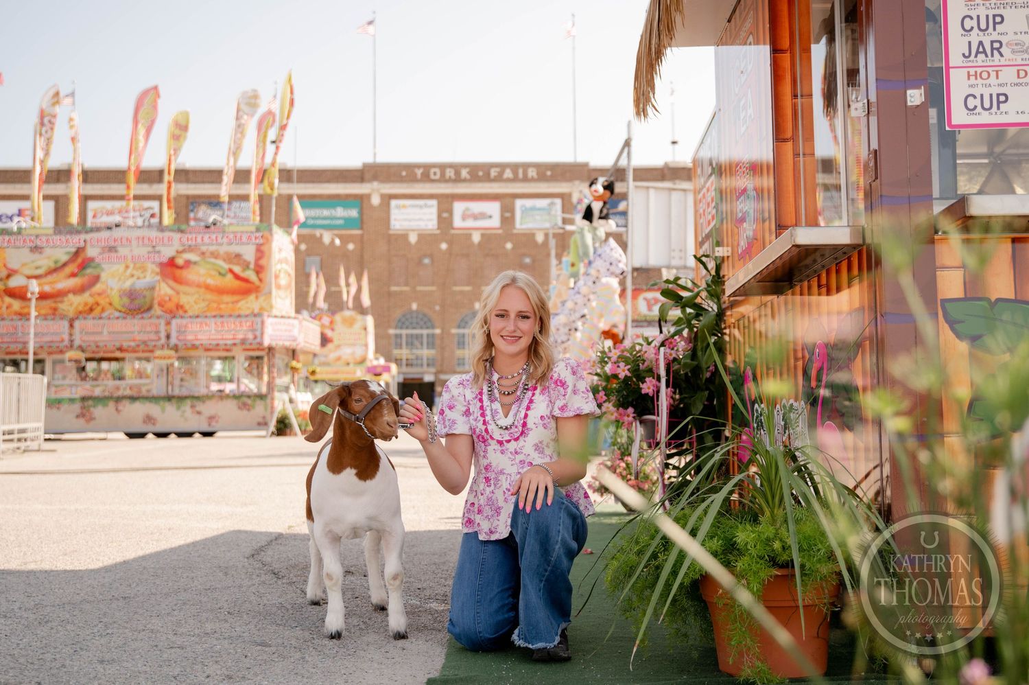 Woman kneeling with a goat at a colorful state fair, wearing a floral top and jeans near food vendor stands.