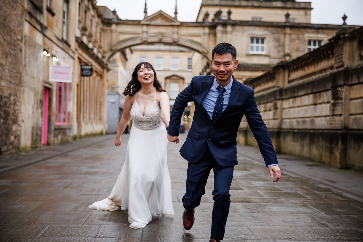 A joyful couple in wedding attire runs hand-in-hand through historic Oxford streets, bride in white gown, groom in navy suit.