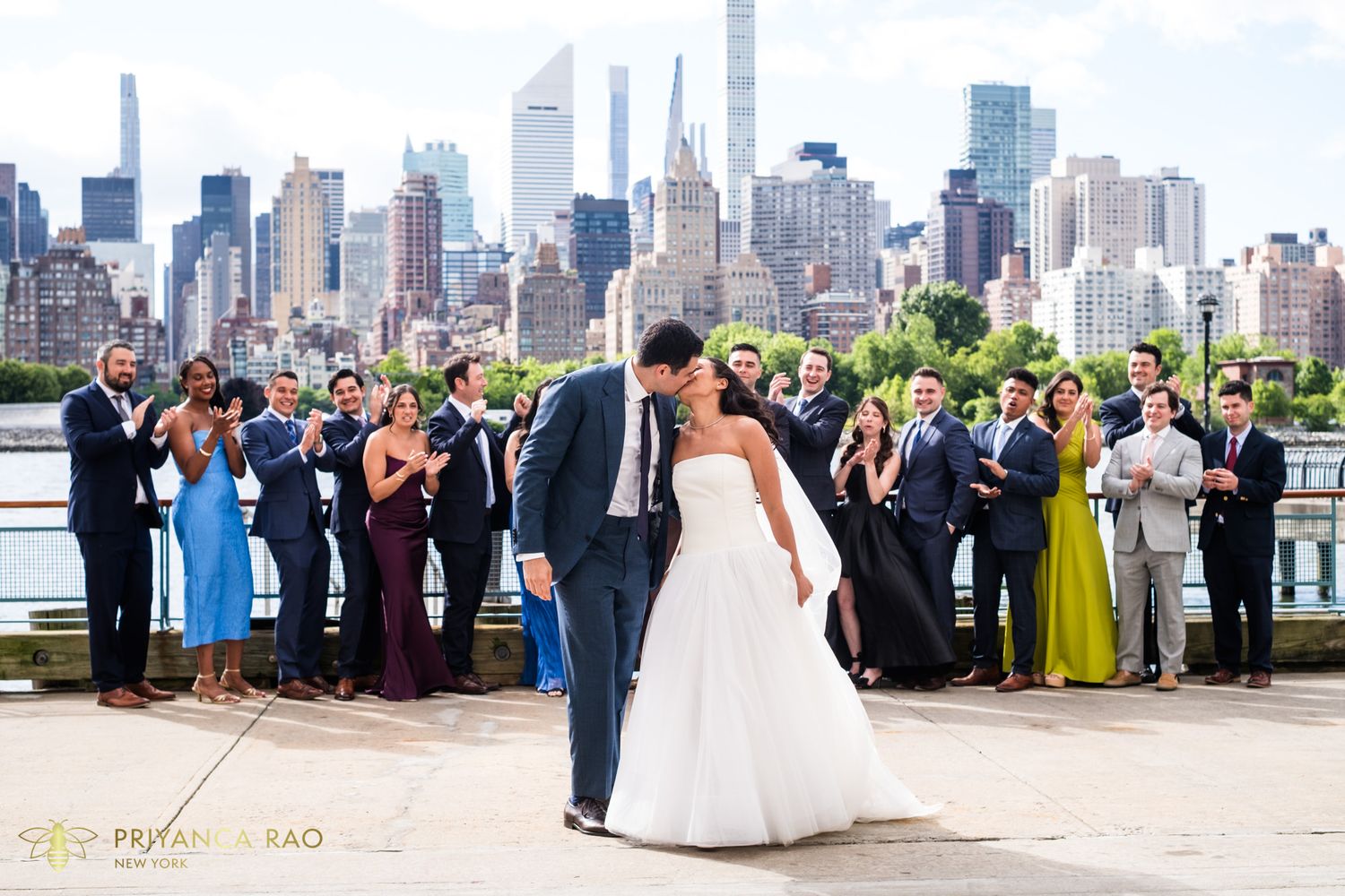 Bride and groom share a kiss with NYC skyline backdrop, surrounded by cheering wedding party on waterfront terrace.