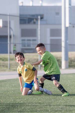 Two young boys in soccer uniforms laugh and play together on a green turf field during a youth soccer match.