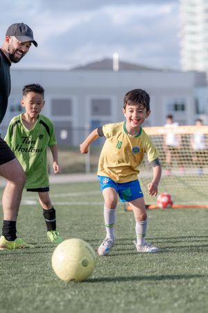 Smiling young boy in yellow jersey dribbling soccer ball during youth league game on turf field.