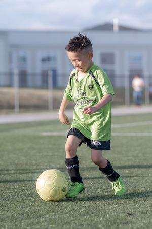 Young boy in green Tim Hortons jersey dribbling yellow soccer ball on turf field.
