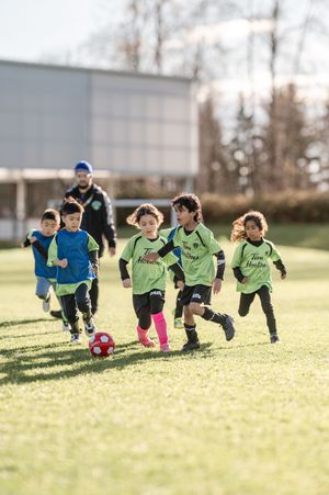Young children in green Tim Hortons jerseys play soccer on a grass field, chasing a red ball on a sunny day.