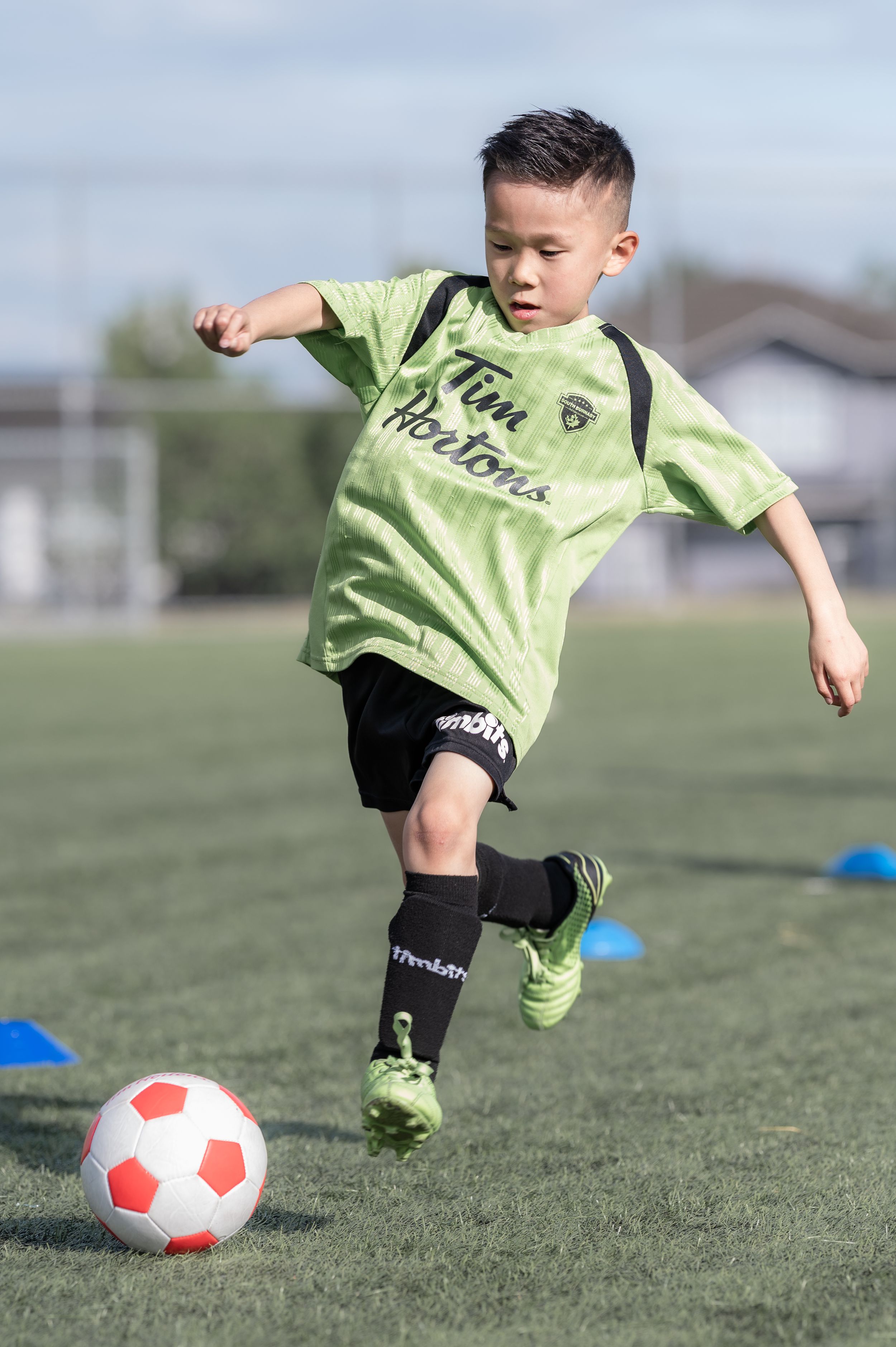 Young boy in Tim Hortons soccer jersey kicking a ball on a green field during youth soccer practice.