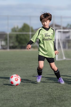 Young boy in green Tim Hortons soccer jersey dribbling a red and white ball on a grass field.