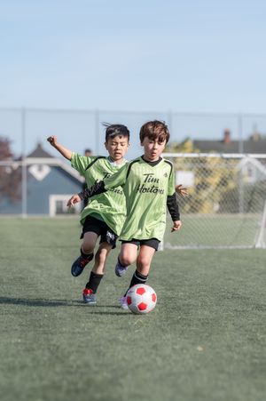 Two young boys in Tim Hortons green jerseys compete for a soccer ball on a turf field.