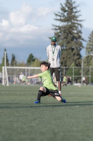 Young boy in green jersey makes dynamic move on soccer field with coach watching in background.