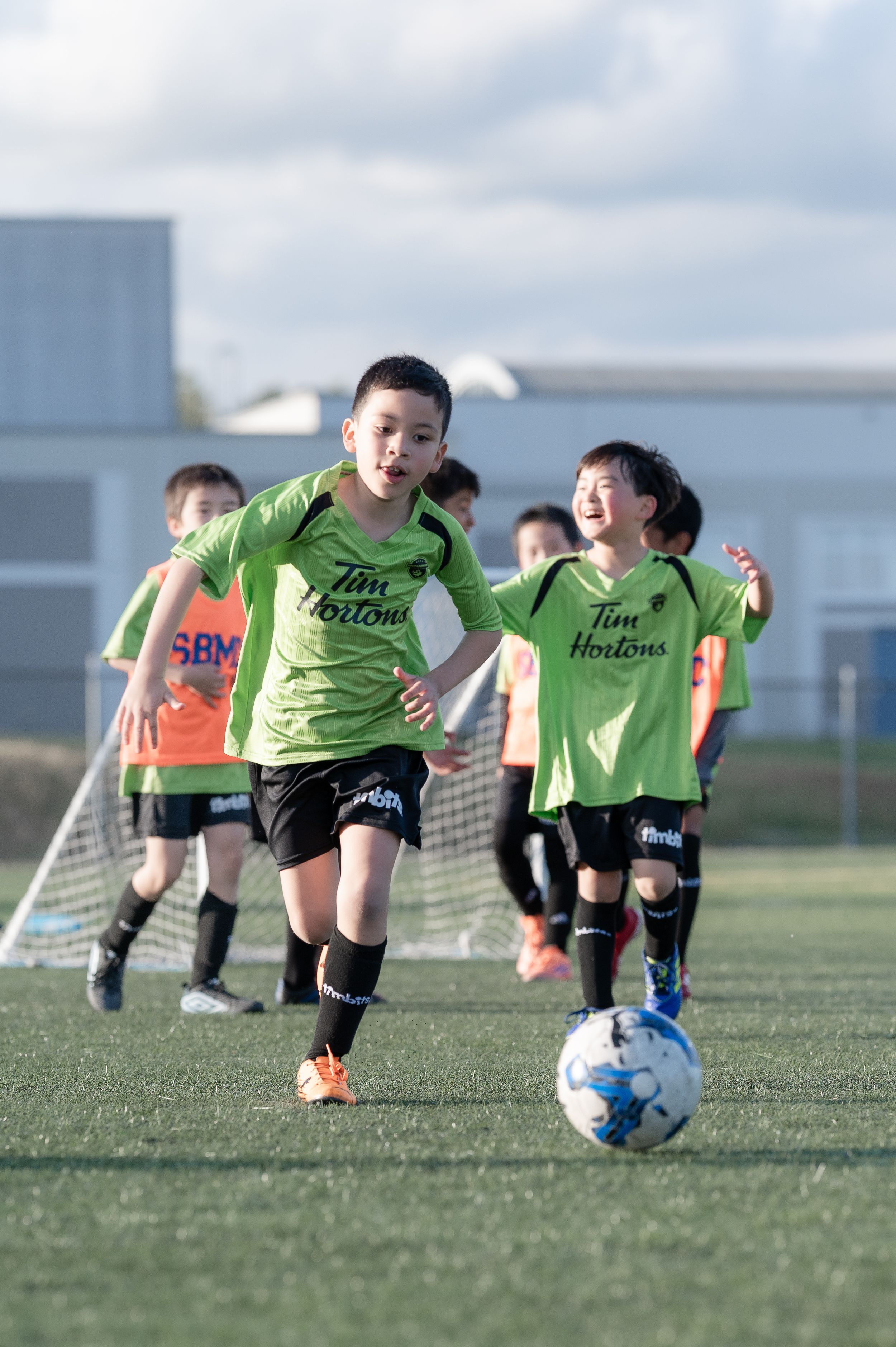 Young kids in green Tim Hortons jerseys play soccer on a turf field during a youth league match.