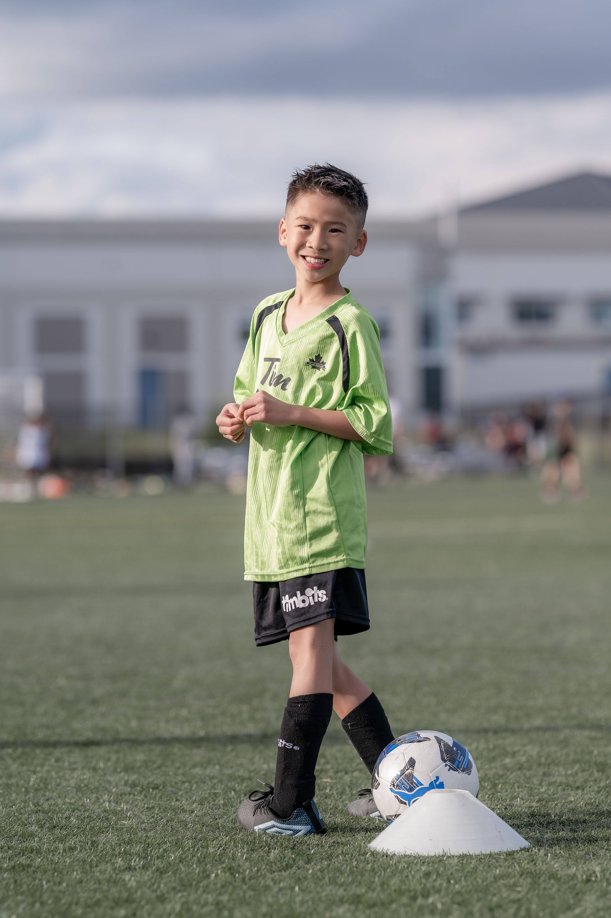 Smiling young boy in green soccer jersey and black shorts standing on field with soccer ball and cone.