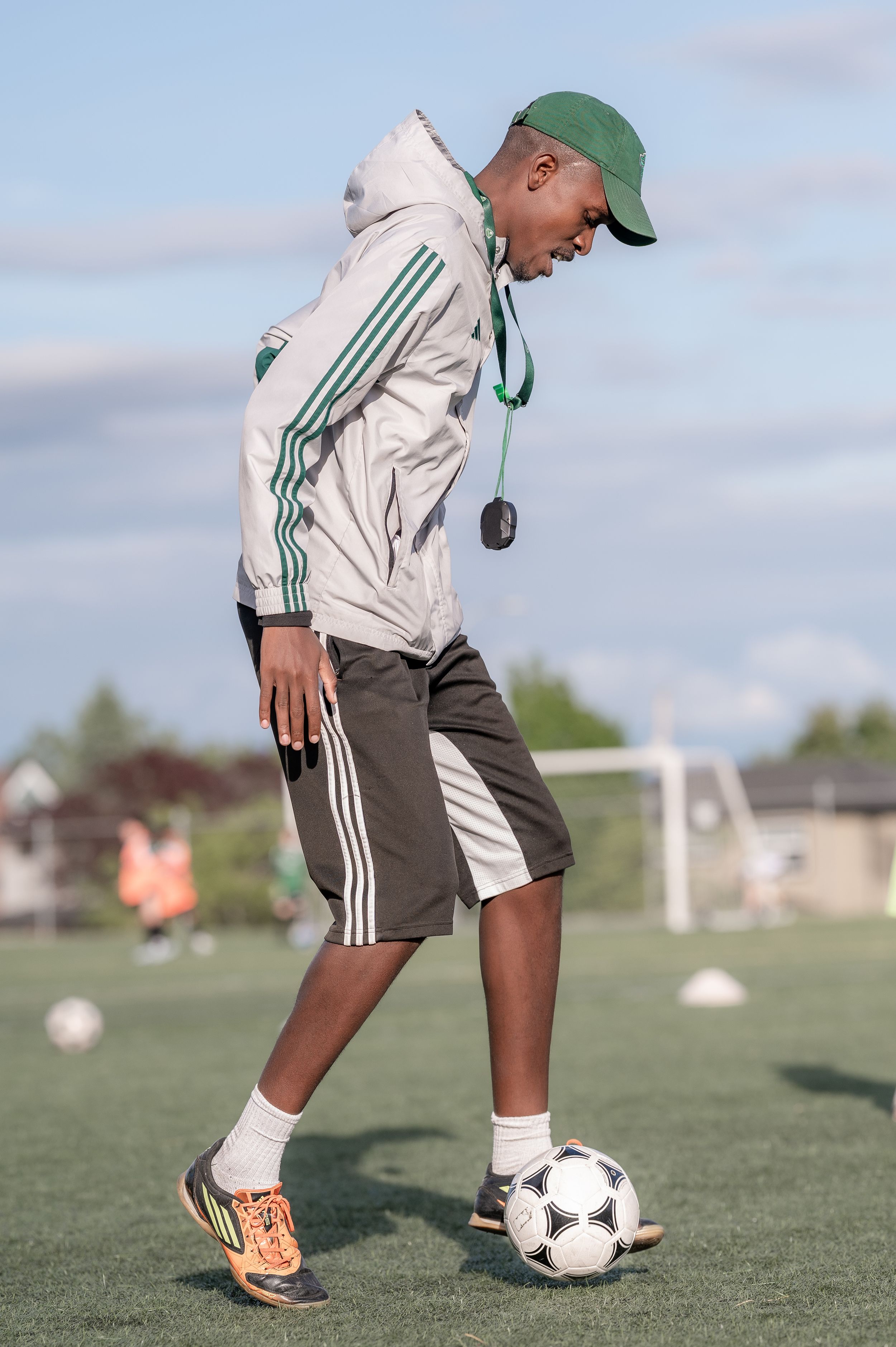 A young soccer coach in a green cap and tracksuit jacket practices ball control on a green field.