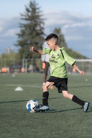 A young boy in a green Tim Hortons jersey kicks a soccer ball during practice on a turf field.
