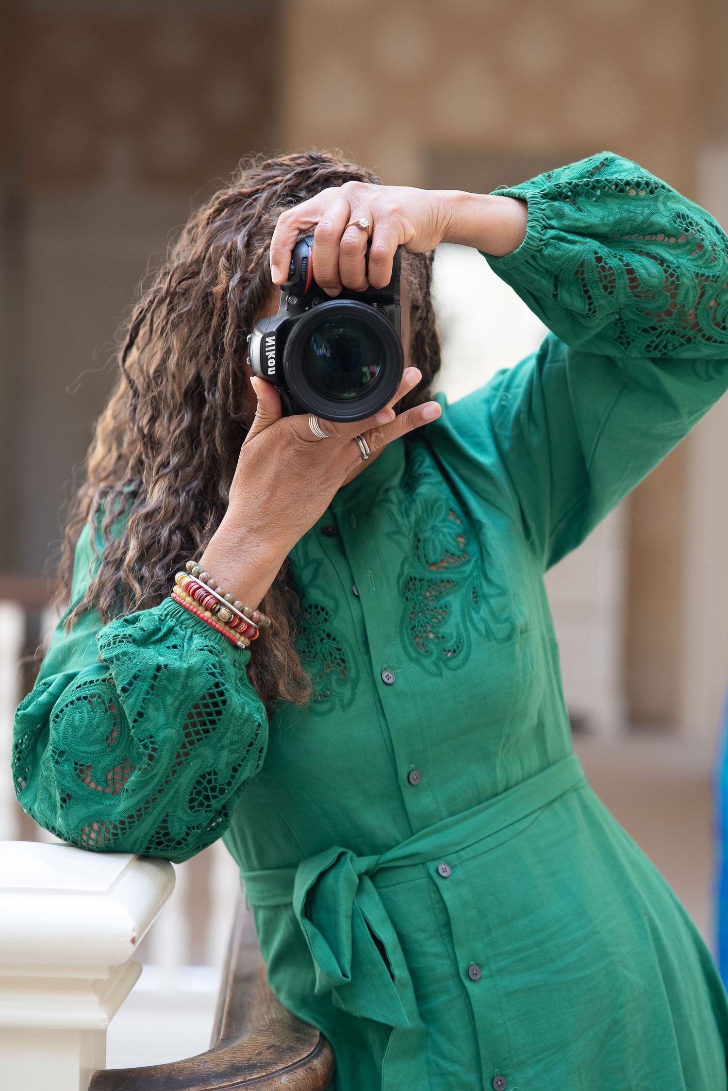Woman in green lace dress photographing with DSLR camera, curly hair, colorful bracelets, outdoor setting.