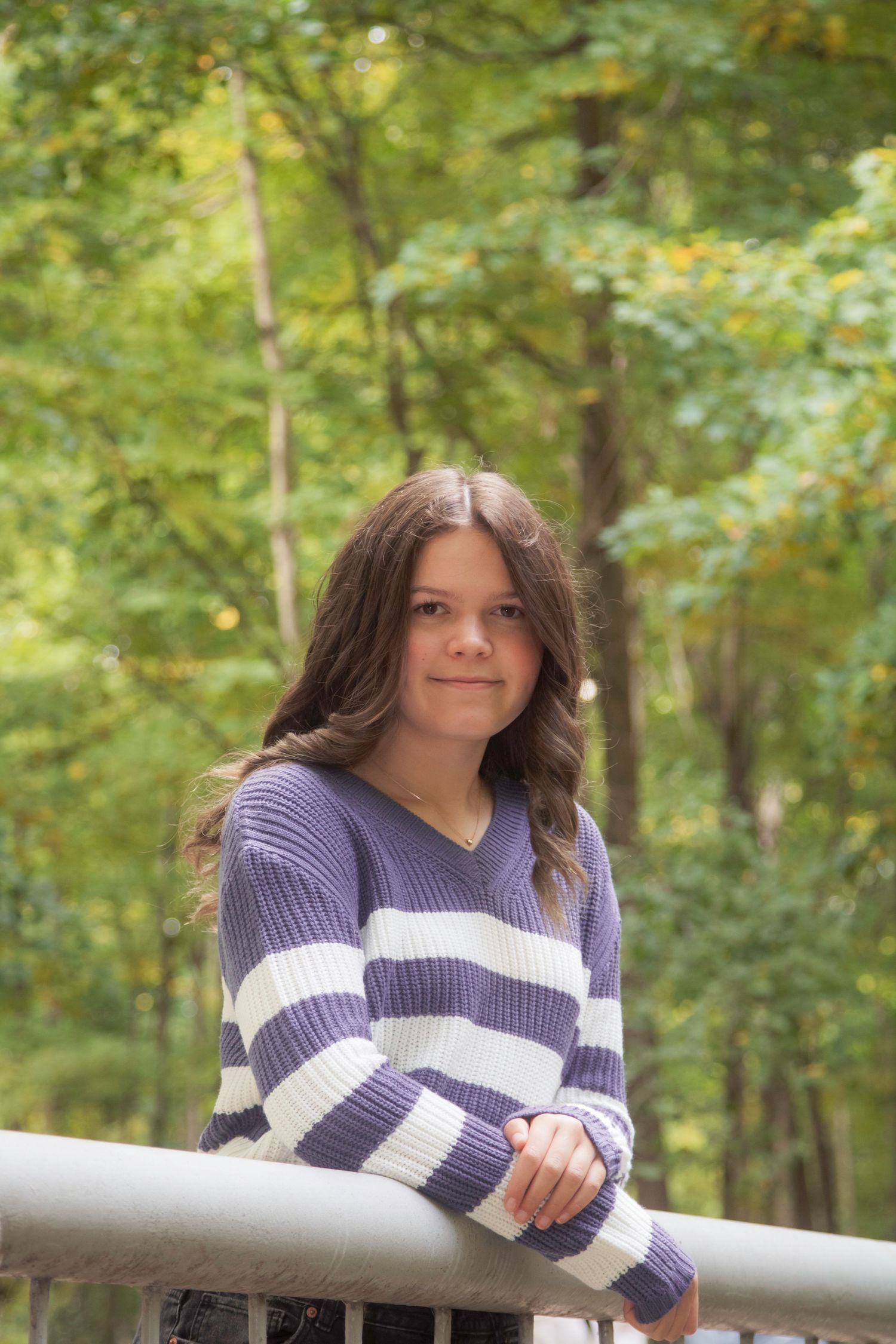 A young girl with long brown hair leans on a metal railing, wearing a purple striped sweater, with autumn trees behind her.