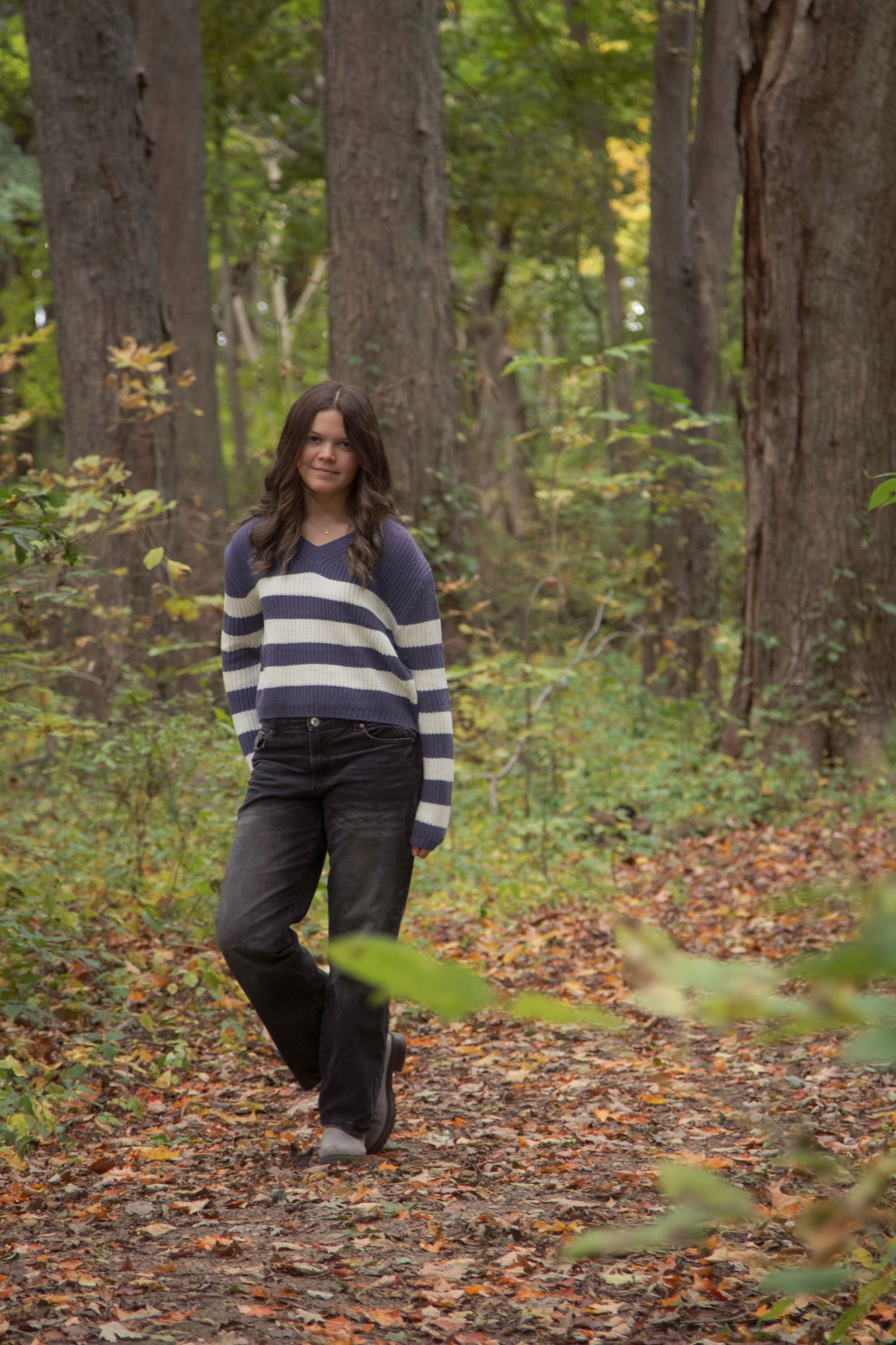 Young woman in striped sweater and black jeans standing on a leaf-covered forest path in autumn.