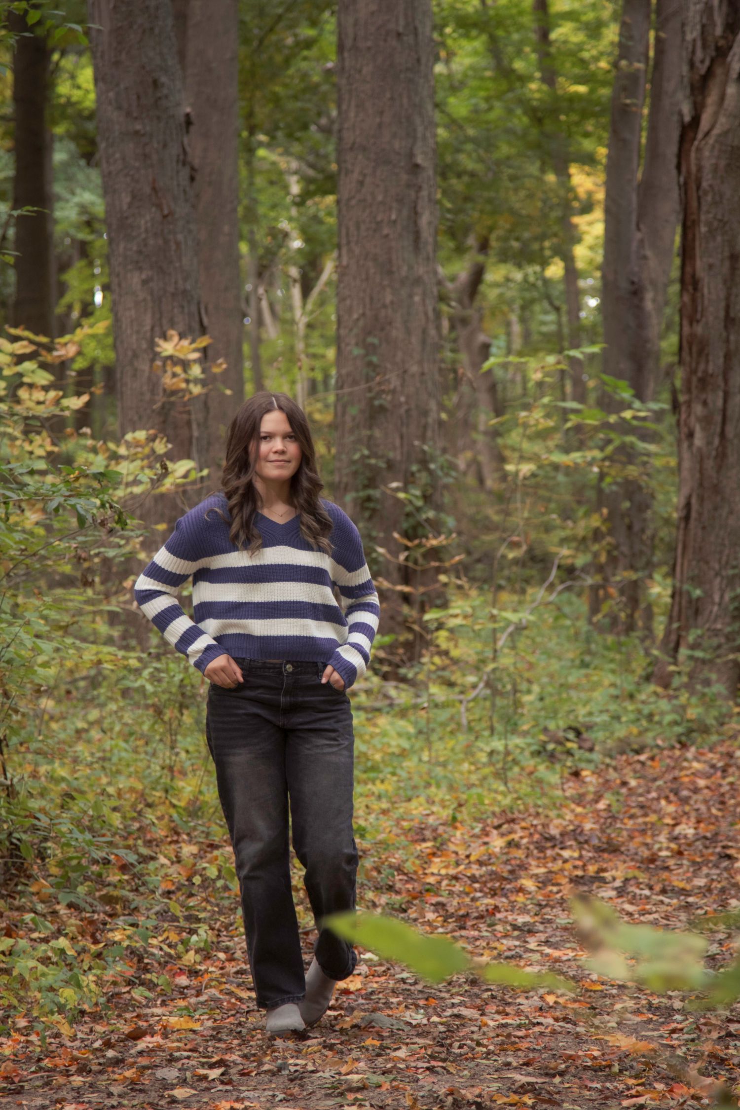 Woman in striped sweater and black jeans walking on a leaf-covered forest path among tall trees in autumn.