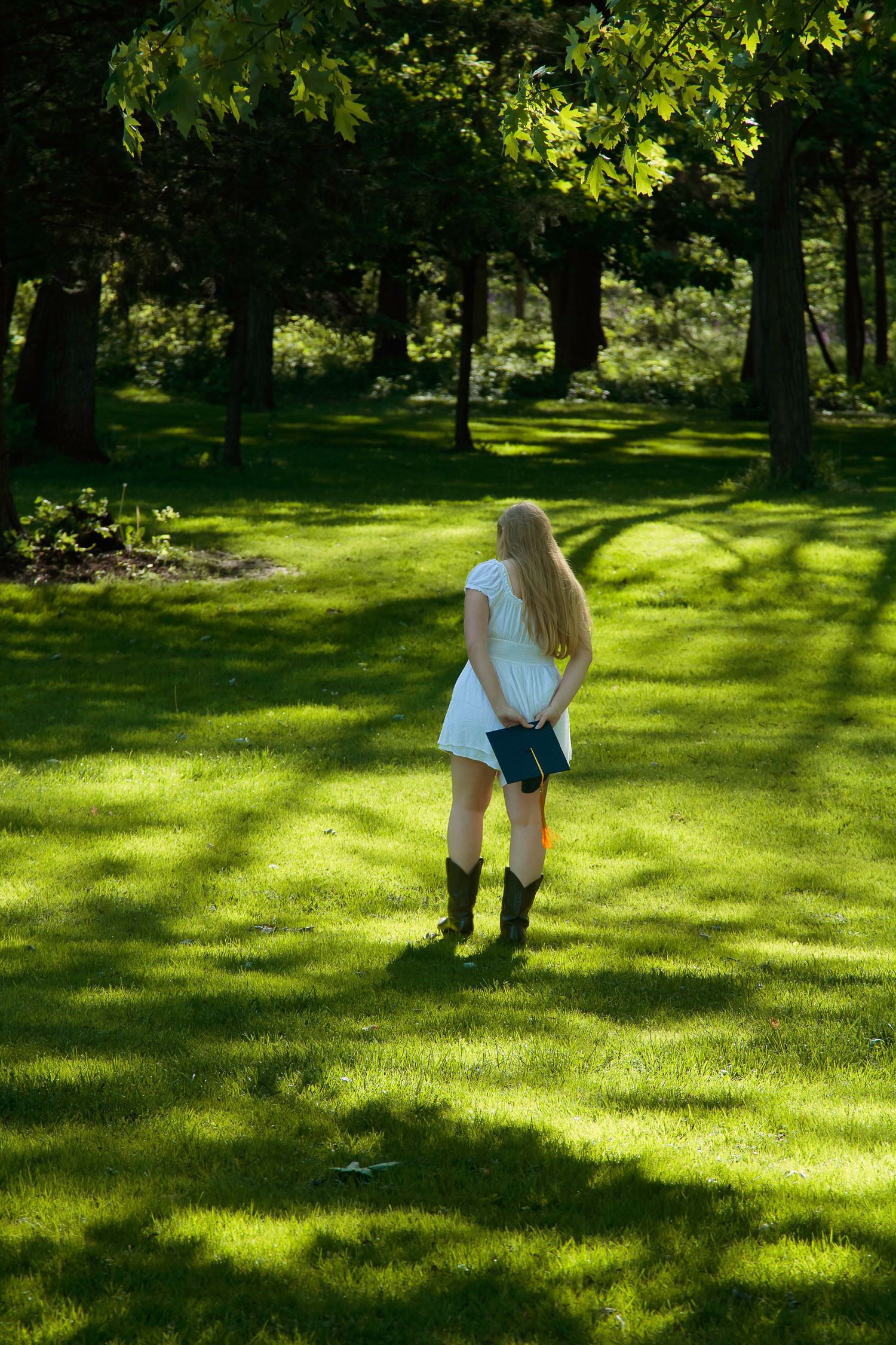Young blonde woman in white dress and cowboy boots holding a book in a sunlit green park.