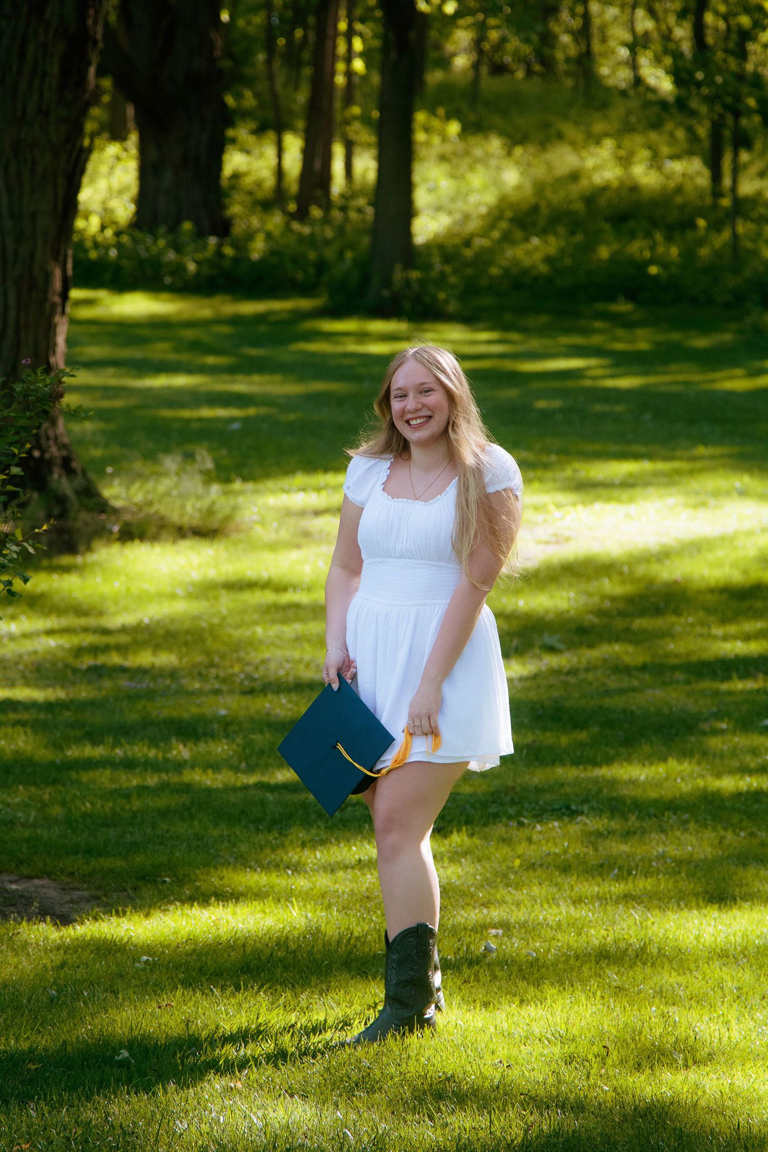 Young woman in white dress holding blue graduation cap smiles outdoors on green lawn with trees.