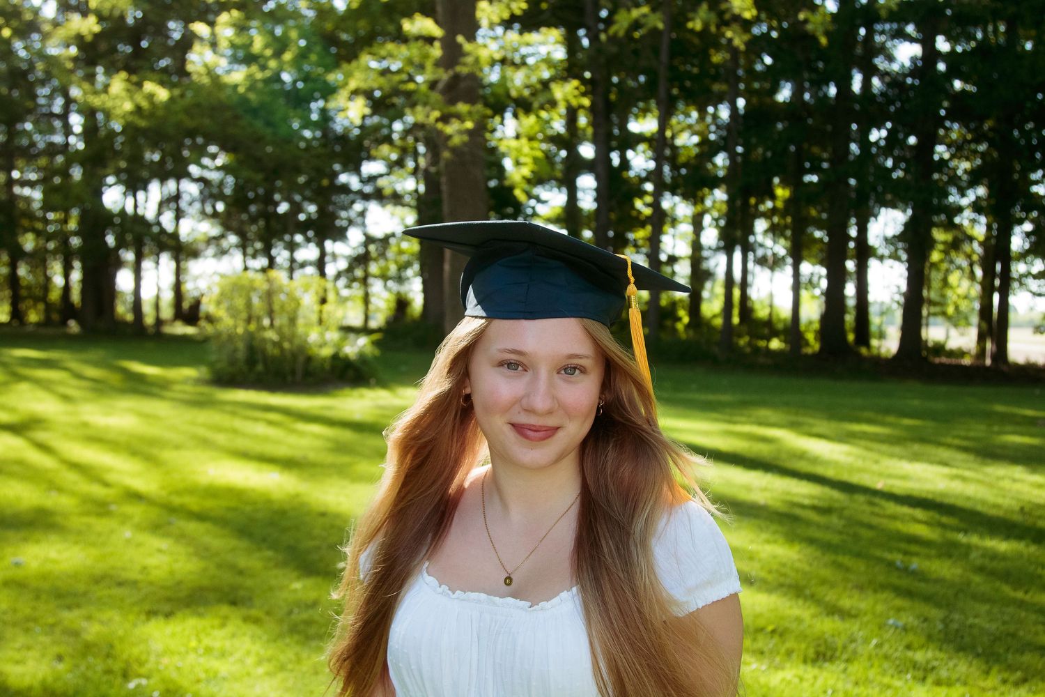 Young woman in graduation cap smiling outdoors on green lawn with trees in background.