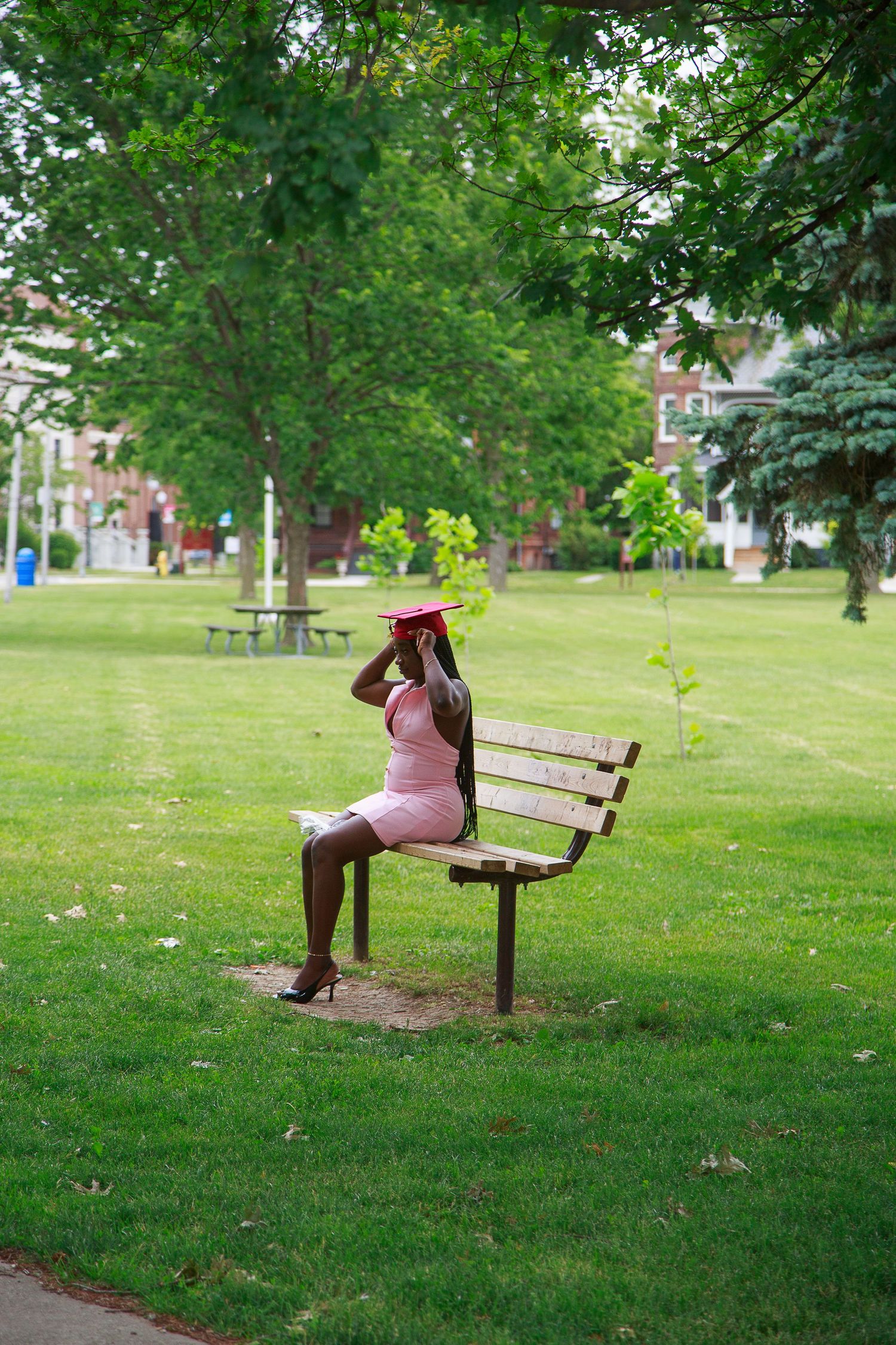 A Black woman in a pink dress and graduation cap sits on a wooden park bench surrounded by lush green trees.