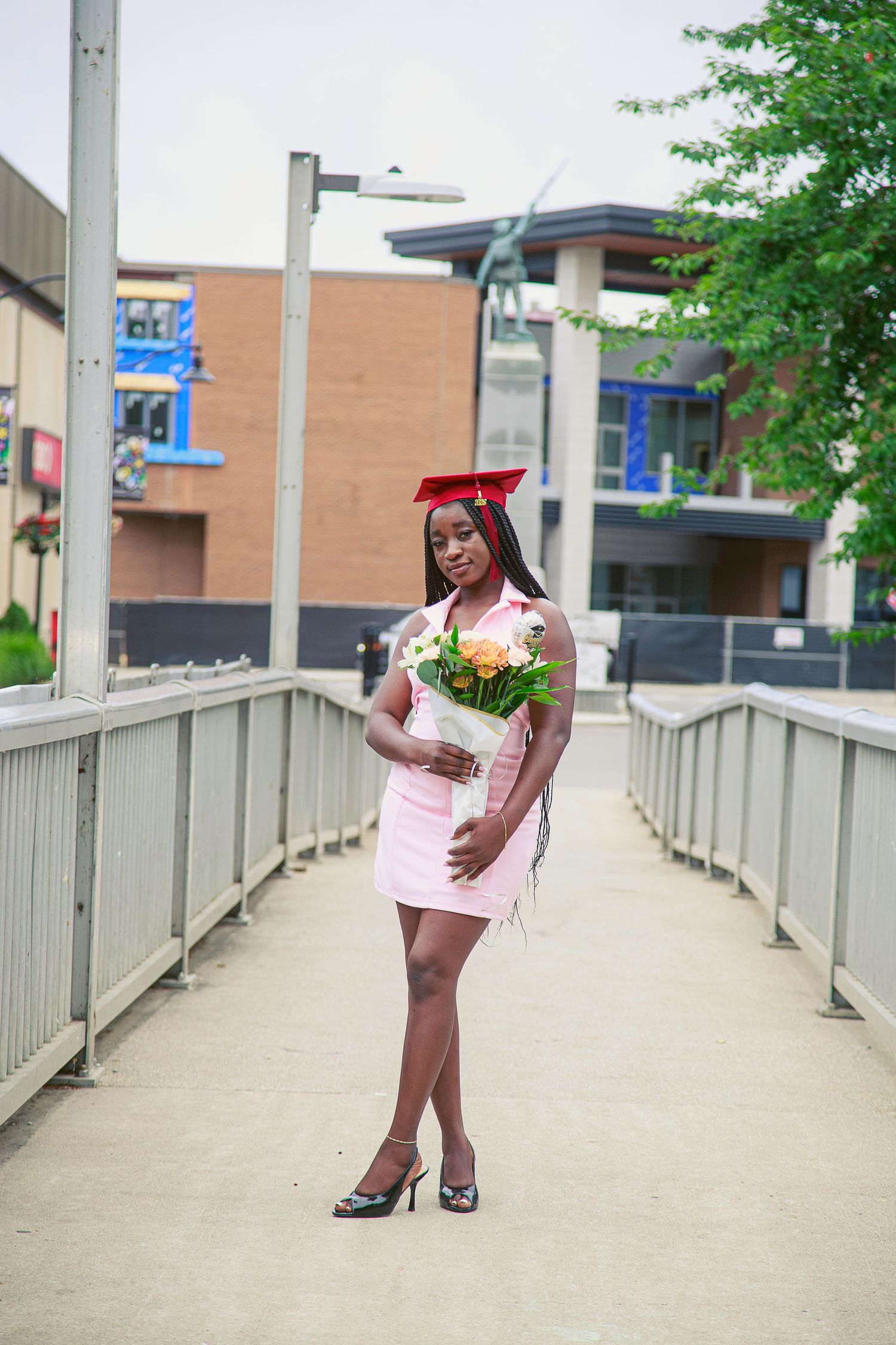 Young woman in pink dress and red graduation cap holds bouquet of flowers on a bridge, celebrating her graduation day.